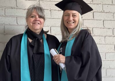 Two women in graduation gowns standing by a white brick wall smiling.