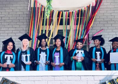 Seven graduates in gowns holding letters spelling TUNITI, with colorful ribbons behind.