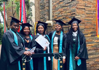 Five female graduates posing outside with certificate and colorful ribbons.