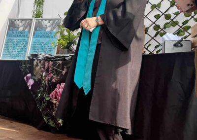 Older graduate speaking on stage in gown near flower display and artwork.