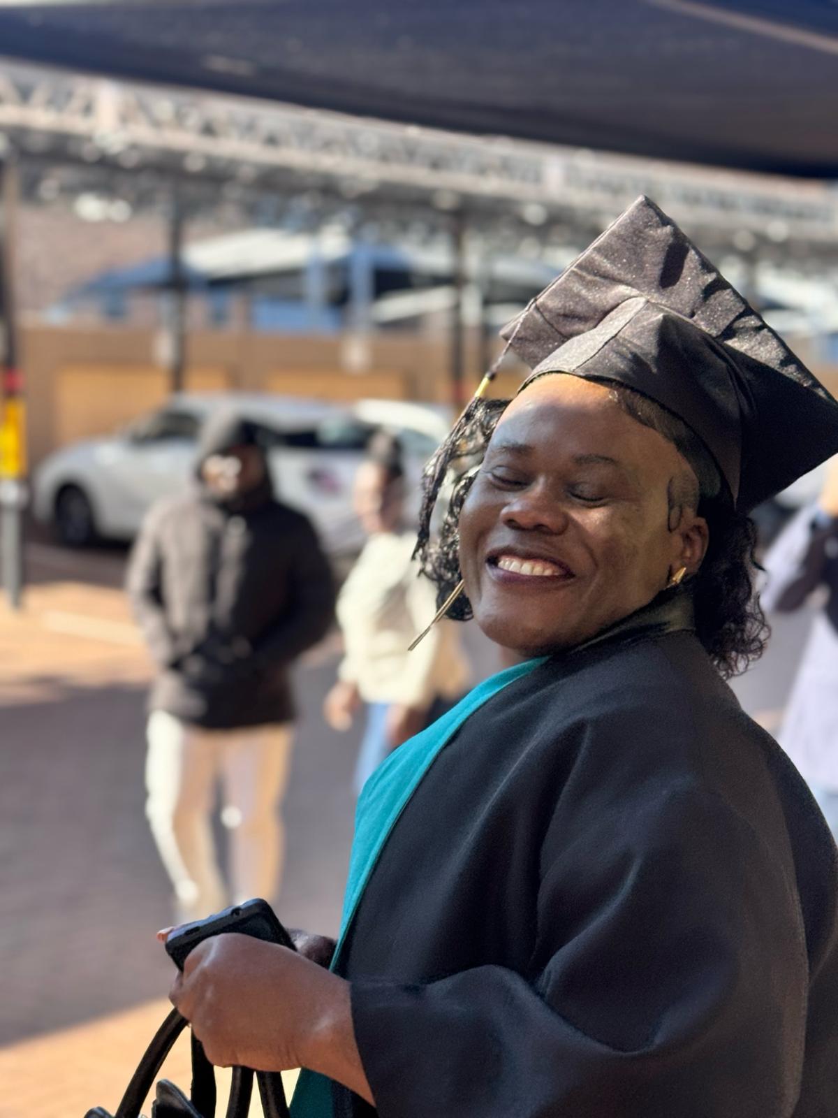 image10 Joyful Tuniti Care Hero graduate in cap and gown beams with pride while holding her bag at the 2025 caregiver graduation event.