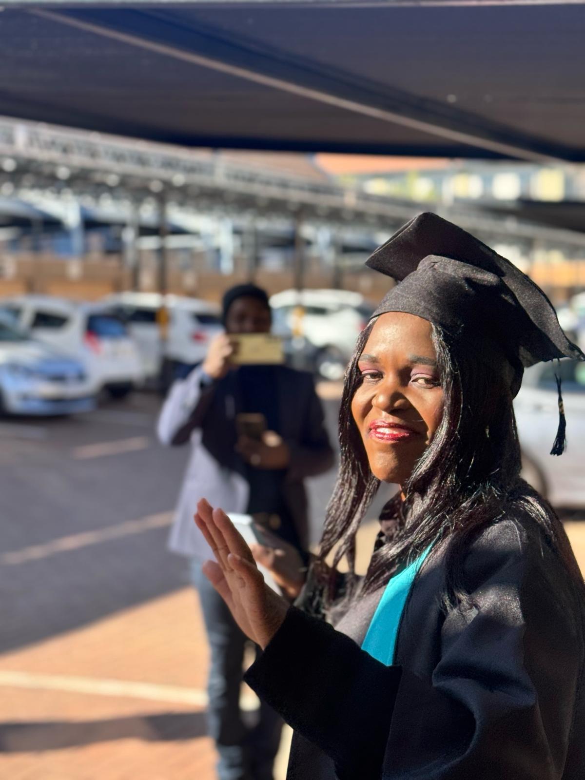 image11 Confident caregiver graduate in cap and gown waves during Tuniti Care Hero graduation celebration, with guest capturing the moment.
