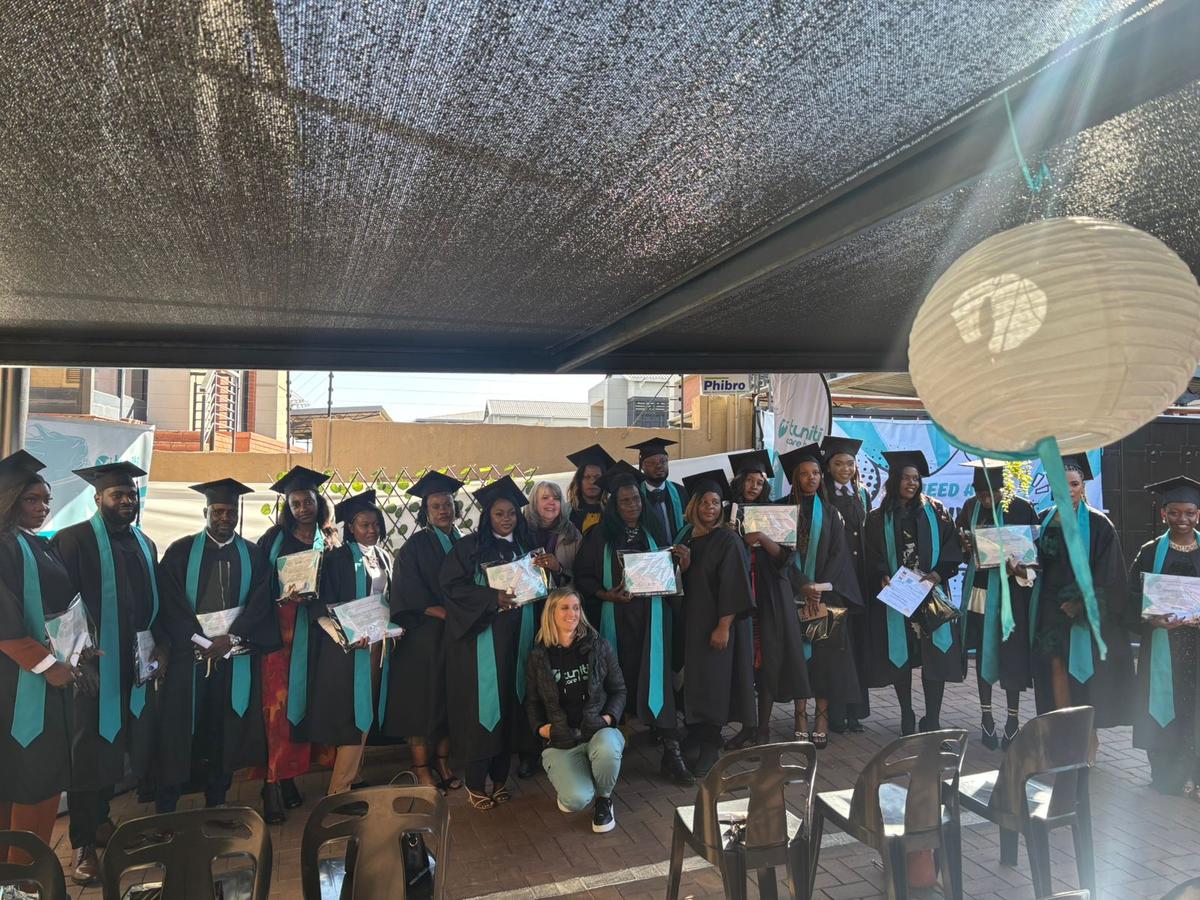 image15 Group of graduates in black gowns and teal sashes celebrating caregiver certification with certificates in hand under decorated canopy at Tuniti Care Hero event.