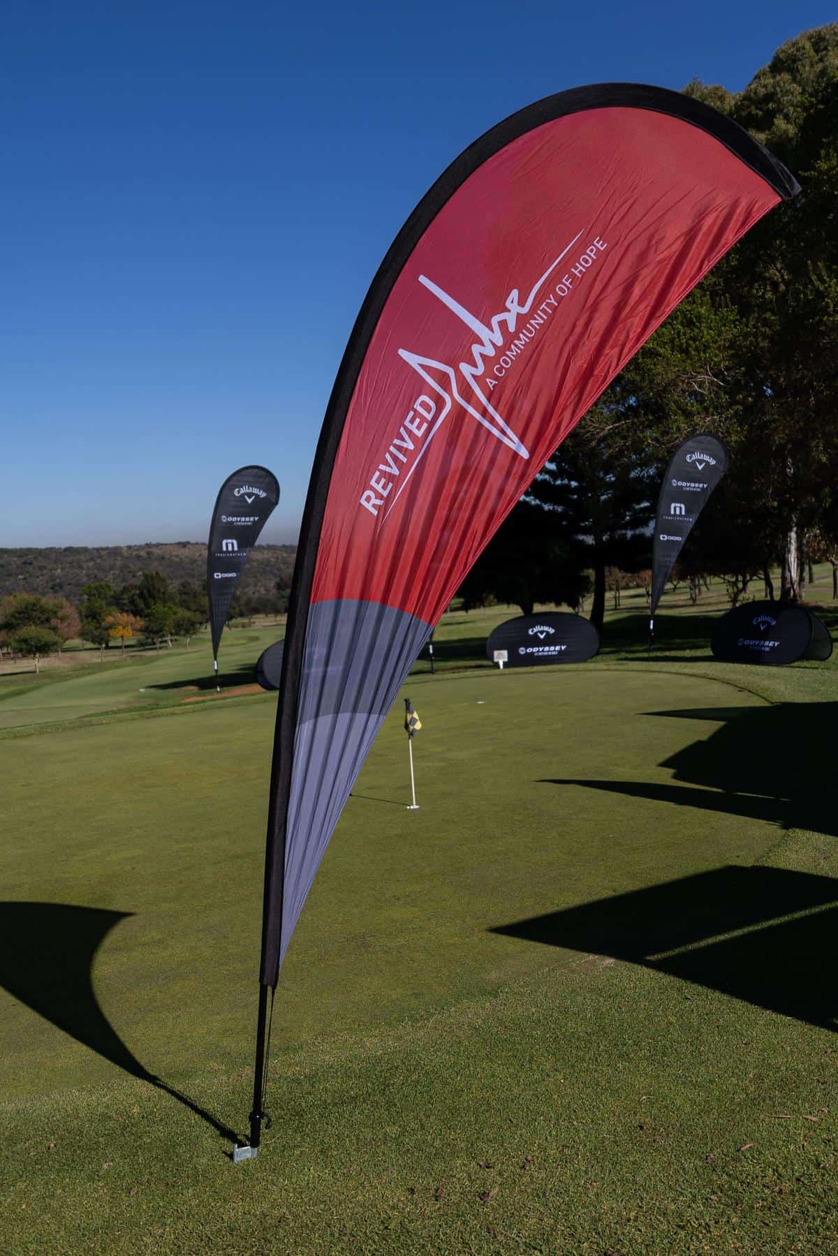 Revived Pulse “A Community of Hope” banner displayed on a golf course during a sunny charity event, surrounded by Callaway and Odyssey sponsor flags.
